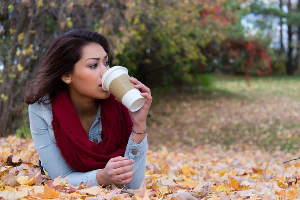 Stilvolle Frau trinkt Kaffee im Liegen auf Herbstblättern — Stockfoto