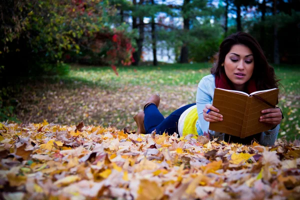 Junge Frau liest Buch im Liegen auf Herbstblättern — Stockfoto