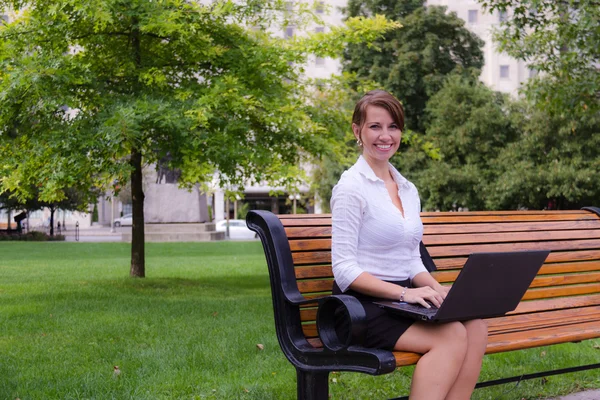Mulher de negócios sorridente no banco do parque que trabalha com computador portátil — Fotografia de Stock