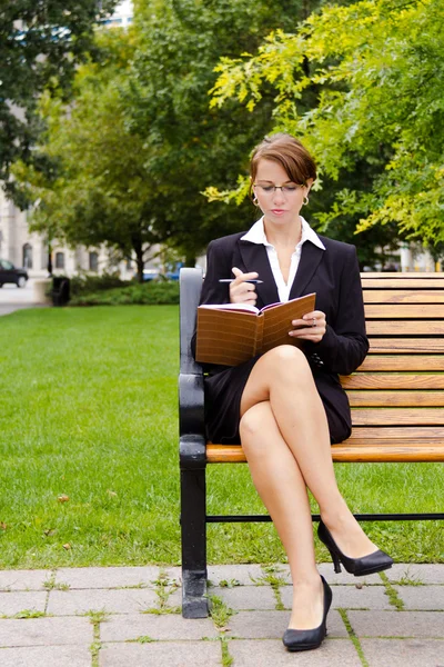 Стильная деловая женщина сидит на скамейке в парке и пишет в блокноте — стоковое фото