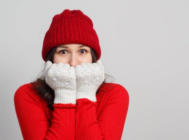 a woman in a red sweater, knitted hat warms her face with mittens. on a white background.