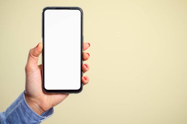 Bir kadının eli dikey olarak, krem arka planda beyaz ekranı olan bir akıllı telefonu tutuyor. Boşluk için.