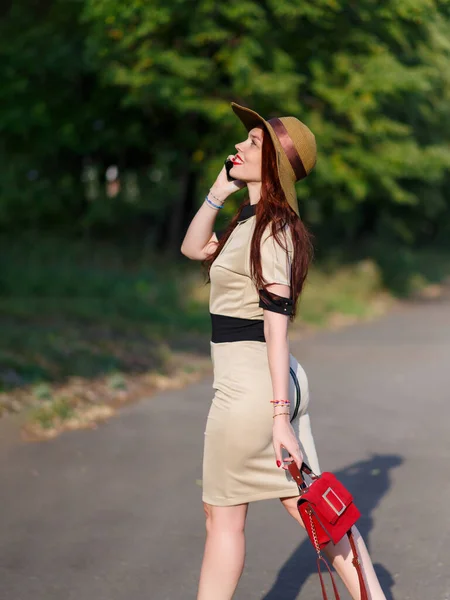 一个戴着宽边帽的快乐的年轻女子提着袋子在公园里散步 还在电话里说着话 — 图库照片