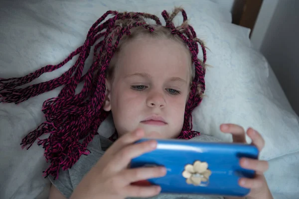 Une fille avec des nattes est couchée au lit et joue avec un téléphone portable. Outils de communication. Le concept de dépendance au téléphone portable — Photo