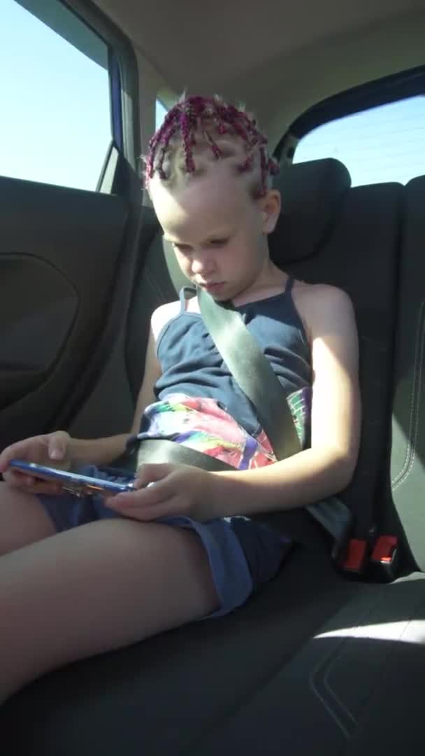 Jenta sitter i baksetet på en bil og leker med telefonen sin. Barnet er lidenskapelig opptatt av spillet. Vertikal video – stockvideo