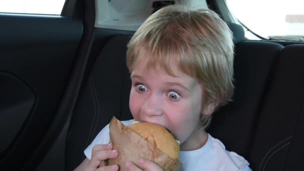 Uma criança come uma sanduíche no carro. A criança está sentada no banco de trás do carro, ele está usando um cinto de segurança. Lanche na estrada — Vídeo de Stock