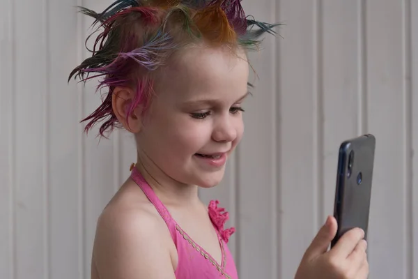 Une belle fille aux cheveux colorés regarde l'écran du smartphone et sourit. Un enfant dans une élégante robe rose — Photo