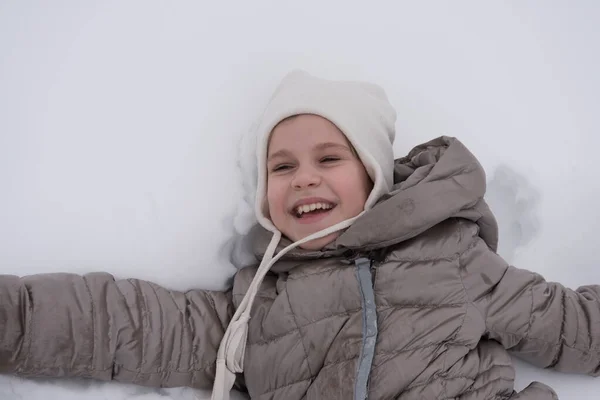 El niño yace en la nieve profunda y sonríe dulcemente. Una chica hermosa en un ambiente blanco como la nieve. Concepto para vacaciones de invierno y paseos. Lugar para el texto —  Fotos de Stock