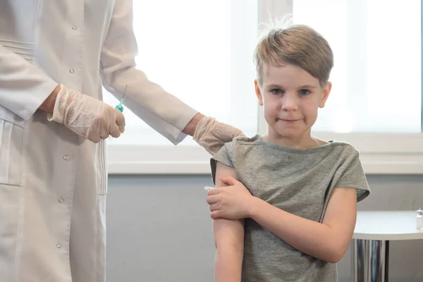 Das Kind lächelt nach der Injektion, nach der Impfung. Der Arzt steht in der Nähe, die Spritze ist sichtbar. Das Kind blickt ruhig in die Kamera und lächelt — Stockfoto