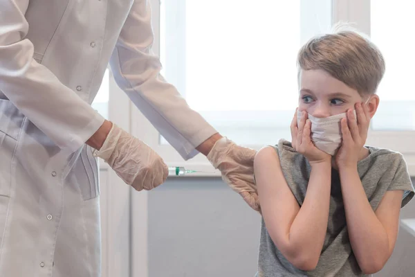 Au premier plan se trouve une seringue dans une main de médecin. En arrière-plan, une image floue d'un enfant dans un masque médical. L'enfant est terrifié à la vue de la seringue. Peur de la vaccination contre le coronavirus — Photo