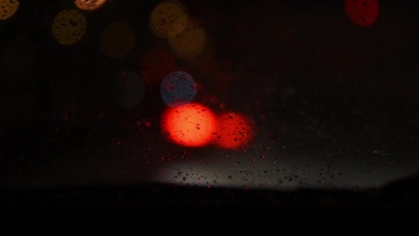 Vista desde el coche hasta las luces de la ciudad nocturna. Coche con parabrisas calentado. El coche está en un atasco de tráfico, la nieve cae sobre el vidrio y se derrite. El limpiaparabrisas está funcionando. Foco suave. centrándose en el vidrio — Vídeos de Stock