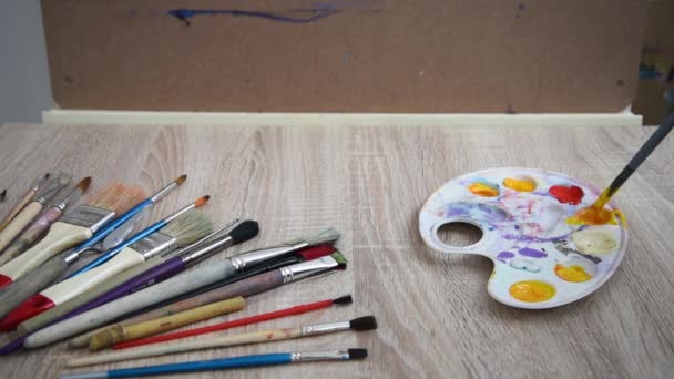O artista mistura tintas em um cavalete para alcançar a cor desejada. Uma variedade de escovas de vários tamanhos e comprimentos são estabelecidas no local de trabalho dos artistas. — Vídeo de Stock