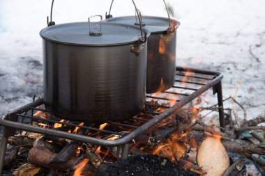 Kış ateşinde yemek pişirmek. İki güzel kamp tavası, altında ateş yakılan bir ızgaranın üzerinde duruyor. Her yerde kar var. Kış turizmi. Maceralar