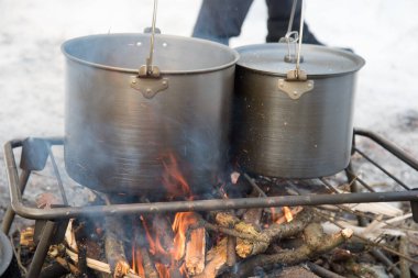 Kış ateşinde yemek pişirmek. İki güzel kamp tavası, altında ateş yakılan bir ızgaranın üzerinde duruyor. Her yerde kar var. Kış turizmi. Maceralar