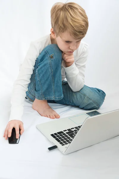 Uma criança do tipo europeu está sentada descalça em frente a um laptop. A loira olha atentamente para o monitor. Ambiente acolhedor em casa. Conceito de ensino em casa. Entusiástico jogo de computador — Fotografia de Stock