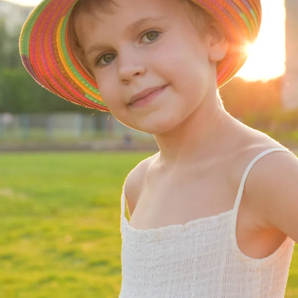 Portrét krásné dívky v letních šatech a barevném klobouku. Dívka se podívá do kamery. Dítě v paprscích zapadajícího slunce — Stock fotografie