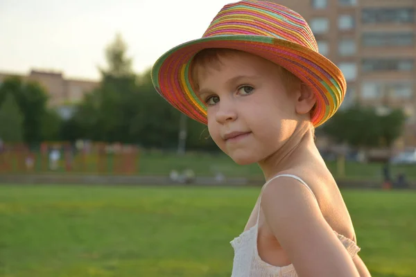 Portrét krásné dívky v letních šatech a barevném klobouku. Dívka se podívá do kamery. Dítě v paprscích zapadajícího slunce — Stock fotografie