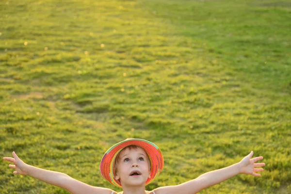 A girl in a multi-colored hat holds her hands above her head. Happy child in the rays of the sun. There is room for text. Green grass background — Stock Photo, Image