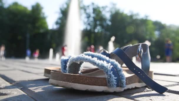 En primer plano están las sandalias ortopédicas para niños. Imagen borrosa en el fondo. Niños divirtiéndose en la fuente. Los chorros de la fuente le permiten refrescarse en los veranos calurosos. Sol, diversión — Vídeo de stock