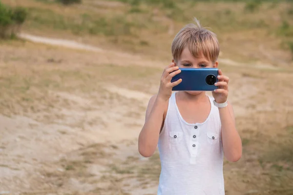The child takes pictures with a smartphone. The phone has 5 cameras. Blonde with short hair plays with the phone. Summer walk in nature with mobile photos — Stock Photo, Image