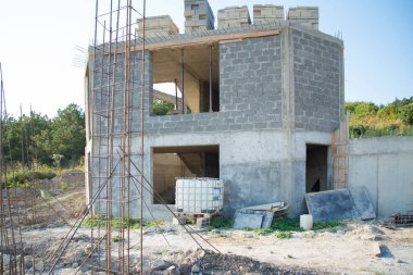 İnşaat alanı. Evin duvarları inşa ediliyor. Yanında betonla dolu bir istinat duvarı var. Metal parçalar görülebilir. Çimento ve beton