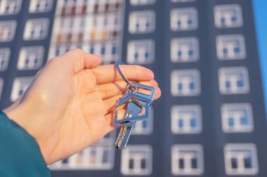 Close-up of key with keychain in shape of house in hand. On the background is multi-storey residential building. Concept of buying and insuring real estate, apartments, mortgages. Copy space.	