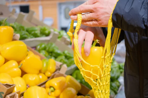 A man\'s hand takes a yellow pepper from a box of peppers in a supermarket and puts it in a string bag. The concept of choosing and buying products in a store, sales, environmental friendliness zero waste.