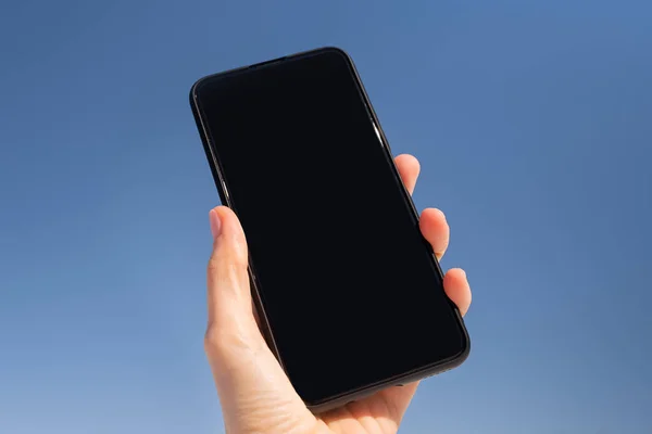 Smartphone with a black screen in a hand against a background of a bright blue sky. Copyspace.