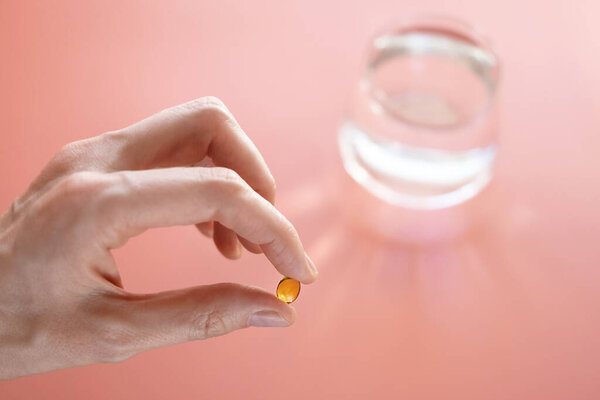 Vitamins of yellow color in a hand on a pink background. Behind - a glass of water. The concept of taking vitamins, dietary supplements, health promotion.