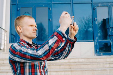 Ekose mavisi kırmızı gömlekli, beyaz, sarışın genç bir adam akıllı telefonuyla panorama fotoğrafı çekiyor. Mobil fotoğrafçılık, video, akıllı telefon kullanımı kavramı.