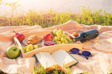 Nehir kenarında piknik. Açık renkli bir yatak örtüsü, içinde meyve, kek ve bir şişe şarap olan ahşap bir tepsi. Batan güneşin ışığı, hoş sakin bir atmosfer, parlak renkler..