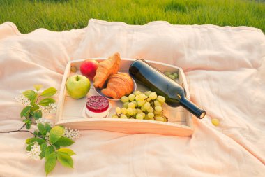Nehir kenarında piknik. Açık renkli bir yatak örtüsü, içinde meyve, kek ve bir şişe şarap olan ahşap bir tepsi. Batan güneşin ışığı, hoş sakin bir atmosfer, parlak renkler..