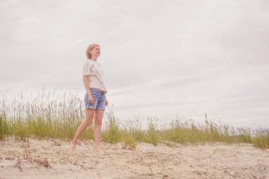 Açık tişörtlü ve mavi kot şortlu beyaz bir kadın nehir kıyısındaki kumun üzerinde duruyor ve uzaklığa bakıyor. Açık hava eğlencesi, rahatlama kavramı..