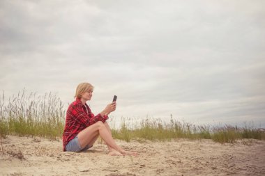 Kırmızı gömlekli ve mavi kot şortlu genç beyaz bir kız nehir kenarındaki kumda oturup akıllı telefona bakar. Açık hava eğlencesi, rahatlama kavramı..