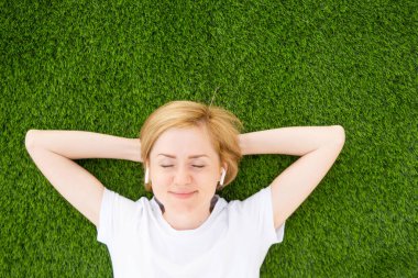 A young Caucasian girl in a white T-shirt and headphones lies on an artificial lawn with closed eyes. The concept of listening to music, podcasts. Minimalism, copy space, top view. clipart