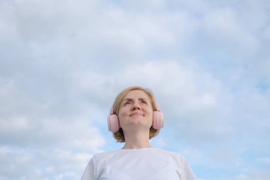 Beyaz tişörtlü ve kulaklıklı genç beyaz bir kız bulutlu mavi bir gökyüzünün arka planında duruyor. Müzik dinleme konsepti, podcastler. Minimalizm, boşluğu kopyala.