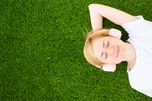 Beyaz tişörtlü ve kulaklıklı genç beyaz bir kız suni bir çimenlikte gözleri kapalı yatıyor. Müzik dinleme konsepti, podcastler. Minimalizm, kopyalama alanı, üst görünüm.