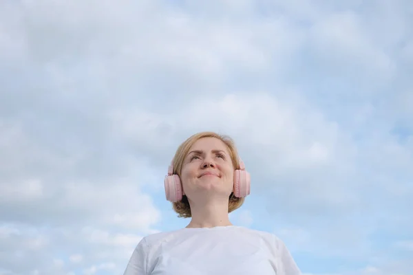 Beyaz tişörtlü ve kulaklıklı genç beyaz bir kız bulutlu mavi bir gökyüzünün arka planında duruyor. Müzik dinleme konsepti, podcastler. Minimalizm, boşluğu kopyala.