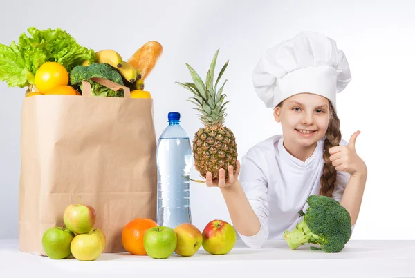 Little happy girl in a cap cook a variety of fresh food. Girl holding pineapple. and shows OK. Positive human emotion, facial expression feeling, attitude. Bag with fresh vegetables and fruits. — Stock Photo, Image