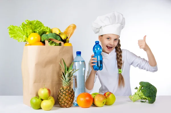 Little happy girl in a cap cook a variety of fresh food. A girl holding a bottle of pure mineral water for cooking healthy meals. A girl holds a finger up. — Stock Photo, Image