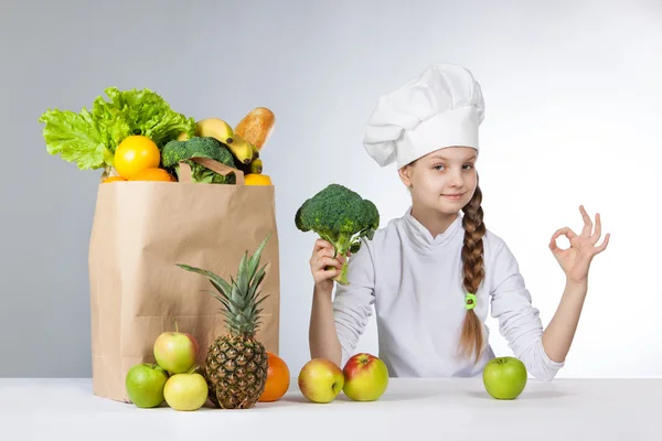 Little happy girl in a cap cook a variety of fresh food. Girl holding broccoli. and shows OK. Positive human emotion, facial expression feeling, attitude. Bag with fresh vegetables and fruits. — Stock Photo, Image