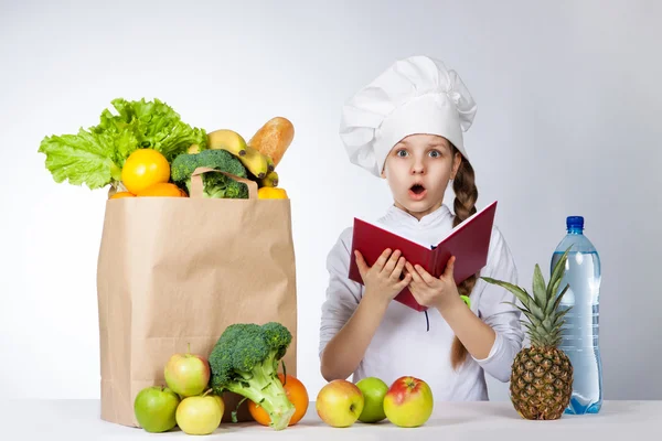 Little girl in a cap cook a variety of fresh food. Girl holds red book in hands. Wonder human emotion, facial expression feeling — Stock Image Little girl in a cap cook a variety of fresh food. Girl holds red book in hands. Wonder human emotion, facial expression feeling Stock Image
