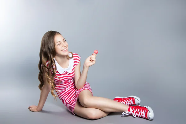 Hermosa chica con una piruleta en la mano está posando sobre un fondo gris. chica en un vestido en rojo con rayas blancas. gusto de moda — Foto de Stock