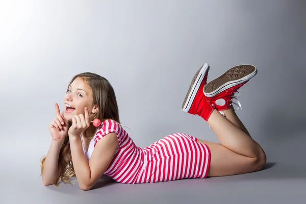 Hermosa chica con una piruleta en la mano está posando sobre un fondo gris. chica en un vestido en rojo con rayas blancas. gusto de moda — Foto de Stock