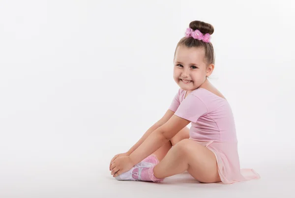 Kleine fröhliche Turnerin, die auf dem Boden sitzt und sein Bein hält. lächelt das Mädchen. heller Hintergrund und Platz für Text — Stockfoto