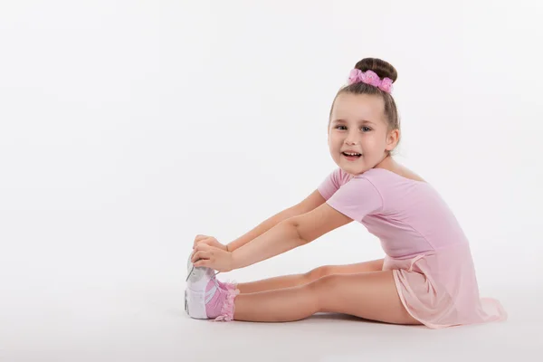 Kleine fröhliche Turnerin, die auf dem Boden sitzt und sein Bein hält. lächelt das Mädchen. heller Hintergrund und Platz für Text — Stockfoto