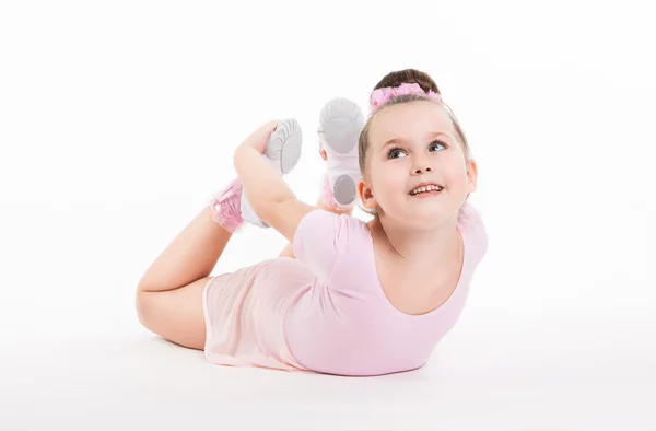 Pequeña chica feliz gimnasta realiza un ejercicio en el suelo sosteniendo su pierna. La chica sonríe. Entrenamiento flexible del gimnasio del traje del niño . —  Fotos de Stock