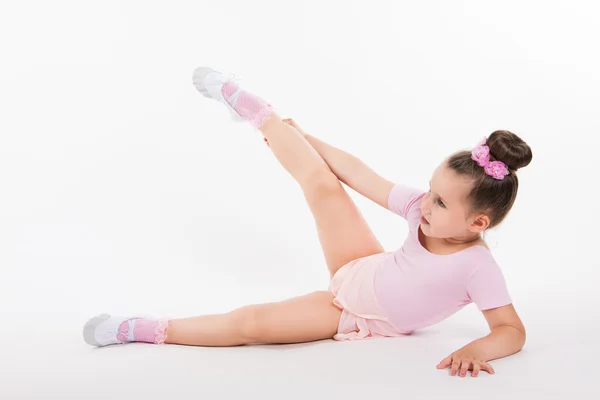 Μικρό κορίτσι εκτέλεση άσκησης κάθεται στο πάτωμα. Αρκετά αστείο γυμναστής σε ένα ροζ φόρεμα σε ανοιχτόχρωμο φόντο. Το παιδί παίρνει ρυθμό — Φωτογραφία Αρχείου