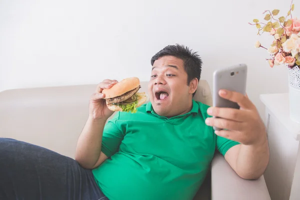 Obese person eating hamburger while using mobile phone — Stock Photo, Image