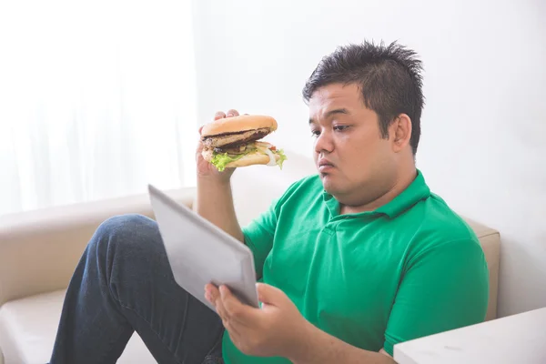 Fettleibige Person isst Hamburger, während sie Tablet-PC benutzt — Stockfoto